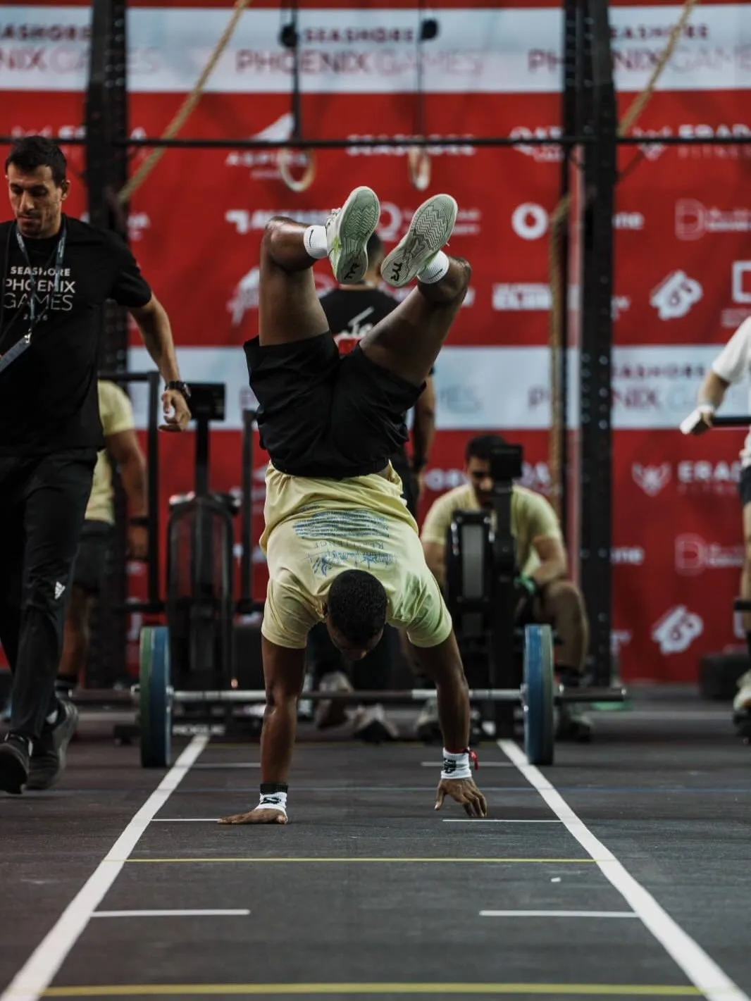 Athlete in a pale yellow T-shirt walking on his hands down a marked lane, coach and rig behind him