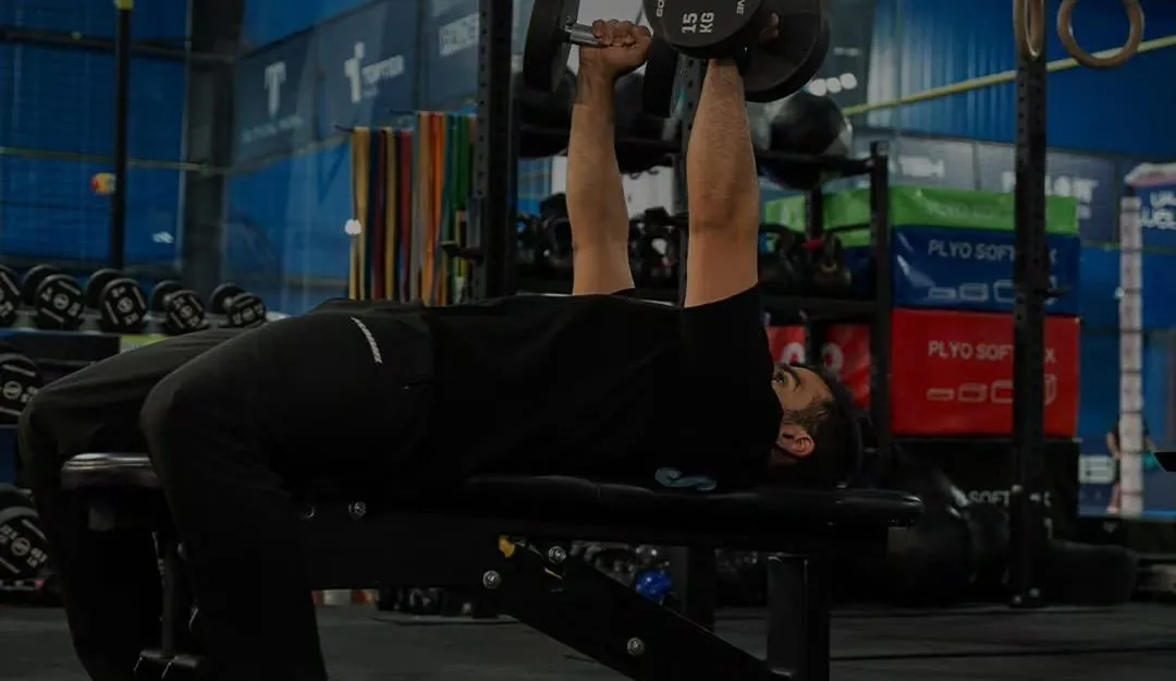 Member pressing dumbbells on a bench, with rings, kettlebells and plyo boxes behind