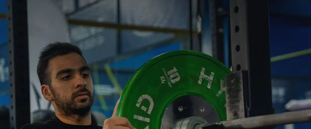 Member in a black SouthFit T-shirt loading a green bumper plate onto a barbell in the rack
